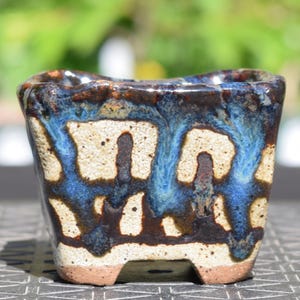 Pode incluir: Um vaso quadrado de cerâmica para bonsai com uma base bege texturizada e salpicada. A borda superior e as laterais apresentam um esmalte brilhante azul vibrante e castanho escuro que escorre para baixo em estrias verticais contrastantes.