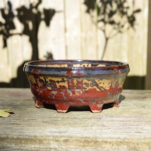 May include: A round ceramic bonsai pot featuring a multi-colored glaze with deep red, earthy brown, and dark blue tones. The pot has a wide, flared rim, a textured surface finish, and is supported by four short, sturdy feet at the base.