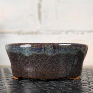 Peut inclure: Un petit pot à bonsaï en céramique brun foncé de forme évasée. Le pot a une texture tachetée et un glaçage brillant et irisé avec des teintes bleues, vertes et violettes. Il repose sur trois petits pieds.