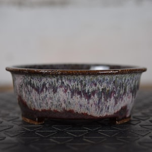 Peut inclure: Petit pot à bonsaï rond en céramique, avec un bord et une base brun foncé. L'extérieur du pot présente un glaçage moucheté dans des tons de blanc, de violet et de bleu. Le pot est conçu avec de petits pieds pour la stabilité.