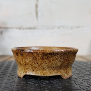 以下が含まれることがあります： 幅が広く浅いデザインの、手作りの小さなセラミックボウル。 ボウルは、茶色とタンの色合いのテクスチャード加工された斑点のある表面が特徴です。 わずかにフレアしたリムがあり、3つの小さな足の上に置かれています。