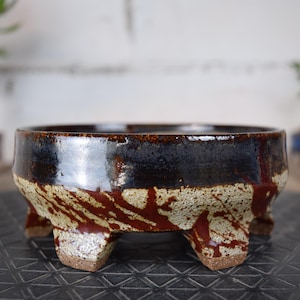 Peut inclure: Pot à bonsaï en céramique fait à la main. Le haut est émaillé brun foncé, la base beige avec des stries rouges. Il possède trois pieds et une forme de bol large et ouvert, idéal pour les petits arbres.
