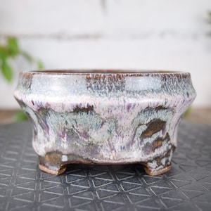 Peut inclure: Un pot à bonsaï en céramique avec un glaçage unique. Le pot a une forme basse et large avec un bord évasé et trois petits pieds. Le glaçage présente des nuances de violet, de vert et de brun, créant un effet marbré. Le pot est conçu pour les bonsaïs.