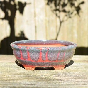 May include: A round ceramic bonsai pot featuring a vibrant orange interior and a textured exterior glaze that blends deep purple and blue tones over a bright orange base, supported by four small, sturdy feet at the bottom of the vessel.