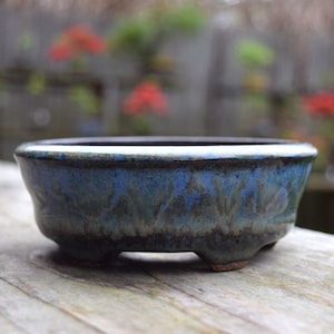 Op de afbeelding: Een kleine, ronde bonsai pot met een blauw en groene geglazuurde afwerking. De keramische pot heeft een laag profiel met drie kleine voetjes. De rand is donker en het glazuur heeft een gevlekt uiterlijk. De pot staat op een houten oppervlak.