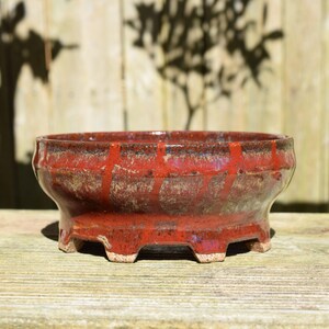 May include: A round ceramic bonsai pot with a glossy red glaze featuring vertical drip patterns and a textured, mottled finish. The vessel has a wide, open top, a slightly tapered body, and stands on four short, sturdy feet with a visible light base.