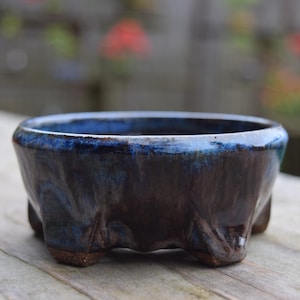 May include: A small, handcrafted ceramic bonsai pot. The pot features a dark brown base with a blue glazed interior and rim. It has a low, wide shape and is supported by three small feet. The background is blurred, suggesting an outdoor setting.