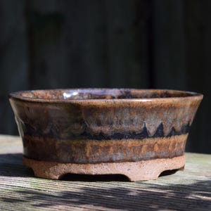 May include: A round ceramic bonsai pot with a flared rim and a footed base. The exterior features a glossy glaze in shades of brown and tan, with a distinct dark horizontal band accented by a subtle drip pattern around the upper section.