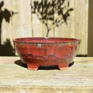 Może przedstawiać: Okrągła ceramiczna doniczka do bonsai z błyszczącą czerwoną glazurą z ciemnymi, nakrapianymi smugami i zaciekami. Naczynie w kształcie misy ma szeroki otwór, gładką krawędź i jest wsparte na czterech krótkich, solidnych nóżkach u podstawy.
