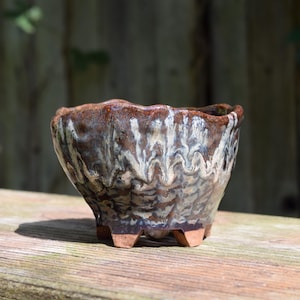 May include: A small, hand-thrown ceramic bowl with a wavy, uneven rim and three short feet. The exterior features a reactive glaze with swirling patterns of dark brown, cream, and grey tones, creating a textured, multi-layered appearance.