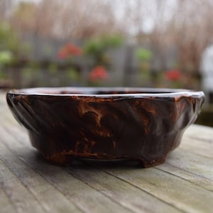 May include: A low, round, brown ceramic bowl with a textured surface. The bowl has a dark brown base with lighter brown marbling. The bowl is sitting on a weathered wooden surface, with a blurred outdoor background.