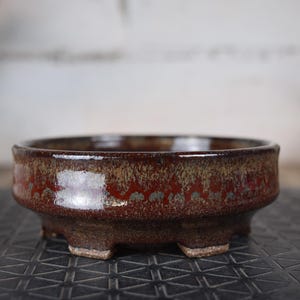Peut inclure: Un pot à bonsaï rond en céramique avec une glaçure marron mouchetée. Le pot a un profil bas, avec un bord légèrement évasé et trois petits pieds. La glaçure présente des nuances de rouge, de marron et des touches de vert.