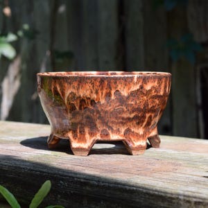 Può includere: Una ciotola in ceramica con finitura lucida caratterizzata da un motivo maculato nei colori marrone scuro, arancione bruciato e crema. La ciotola presenta un'ampia apertura circolare ed è sostenuta da quattro piedini corti e robusti.