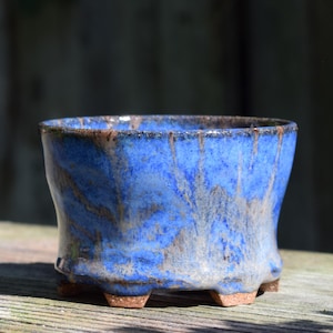 May include: A small, round ceramic planter with a vibrant blue glaze that features mottled brown and tan undertones. The pot has a wide, open top, slightly flared sides, and stands on four small, sturdy feet made of exposed, unglazed brown clay.