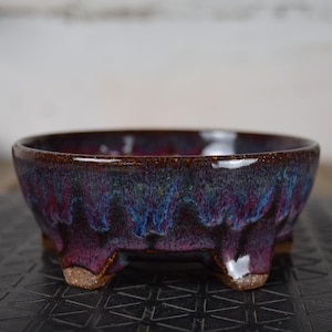 May include: A ceramic bowl with a glossy finish, featuring a blend of purple, blue, and brown hues. The bowl has a rounded shape and is supported by three small feet. The interior is a deep, rich colour.