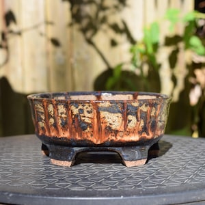 May include: A round ceramic bonsai pot featuring a textured exterior with a mottled tan and black glaze. The surface displays vertical drip patterns in shades of reddish-brown and dark brown. The pot is supported by four short, sturdy feet.