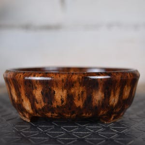 Peut inclure: Petit pot à bonsaï rond en céramique marron, finition brillante. Le pot présente un motif texturé de rayures verticales marron foncé et beige. Il repose sur trois petits pieds et convient aux bonsaïs.