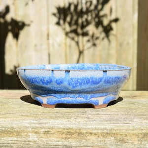 May include: A shallow, oval-shaped ceramic bonsai pot featuring a reactive glaze with shades of deep blue, light blue, and white. The pot has a wide rim, a tapered body, and four small, integrated feet at the base, revealing a natural clay color.