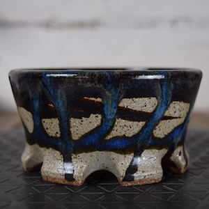 Peut inclure: Un pot de bonsaï en céramique au design unique. Le pot présente un glaçage bleu foncé avec des stries de bleu clair et de beige. Le pot a une large ouverture et une base robuste à trois pieds.