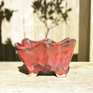 May include: A small, four-legged ceramic planter featuring a glossy, deep red glaze with subtle dark undertones. The rim has a distinct, undulating, wavy shape, and the base shows a small portion of unglazed, light-colored clay at each foot.