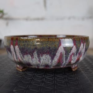 Peut inclure: Un pot de bonsaï en céramique avec une finition émaillée. Le bol est rond avec trois petits pieds. L'émail présente un mélange de couleurs, dont le vert, le violet, le blanc et le rouge. Le pot est conçu pour les bonsaïs.