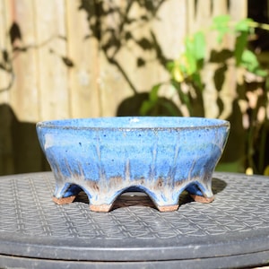Może przedstawiać: Okrągła ceramiczna doniczka do bonsai z żywą niebieską glazurą, która spływa na jaśniejszą, piaskową podstawę. Naczynie ma szeroki otwór i wspiera się na czterech wyraźnych, łukowatych nóżkach, które odsłaniają surową, nieszkliwioną brązową glinę.
