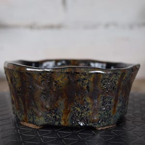Op de afbeelding: Zeshoekige bonsai pot van keramiek met een golvende rand en een donkere, getextureerde glazuur. De pot heeft een gevlekt patroon in bruin, groen en goudtinten. Ontworpen voor bonsai teelt.