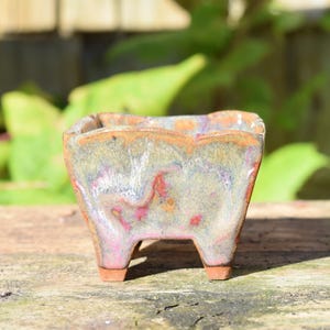 May include: A small, square ceramic planter with four short legs. The glaze features a mottled blend of muted green, soft pink, and earthy brown tones with a glossy finish. The rim has a slightly uneven, organic shape, and the base is unglazed.