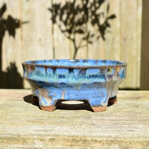 May include: A round ceramic bonsai pot featuring a vibrant blue glaze with dark brown and green drip accents. The pot has a wide, shallow basin, a textured exterior surface, and is supported by four short, sturdy feet with an exposed brown clay base.