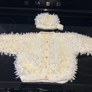 May include: A cream-coloured, hand-knitted baby cardigan and matching hat set. The cardigan has a button-down front and textured, looped detailing. The hat features a similar looped design. The set is displayed on a black surface.