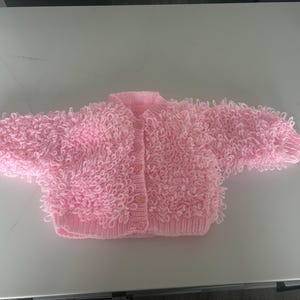 May include: A pale pink knitted baby cardigan with a textured, looped design. The cardigan has long sleeves and a button-up front. The soft, fluffy texture gives the sweater a unique look. The garment is designed for infants and toddlers.