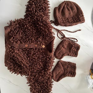 May include: A brown knitted baby outfit set, featuring a hooded vest with button fastenings, a beanie hat with ties, and two pairs of booties. The set is made of a textured, curly yarn.