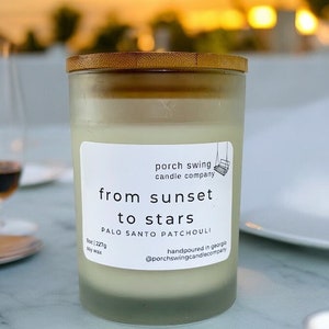 May include: A glass candle jar with a wooden lid. The candle is labelled "porch swing candle company" and "from sunset to stars palo santo patchouli". The candle is handpoured in Georgia and weighs 348g. The candle is made of soy wax.