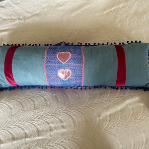 Puede incluir: Una almohada larga azul y rosa con ribete de pompones azules y borlas rosas. La almohada tiene dos corazones rosas con lentejuelas en la parte delantera.