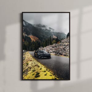 May include: Framed print featuring a black sports car driving on a mountain road. The road winds through a landscape of rocky terrain, evergreen trees, and a cloudy sky. The print is displayed on a white wall.