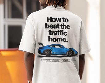May include: White t-shirt with the text "How to beat the traffic home." and a blue sports car graphic. The shirt also includes additional text below the car. The person is wearing a white cap and blue jeans.