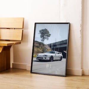 May include: A framed photograph of a white sports car speeding on a road. The car is the primary focus, with a blurred background of a bridge and trees. The frame is black, and the image is leaning against a white wall.