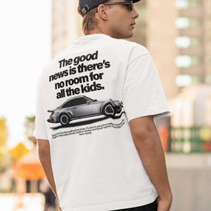 May include: White t-shirt with the text "The good news is there's no room for all the kids." and a silver car graphic. The shirt also has additional text below the car. The person is wearing a black cap and sunglasses.