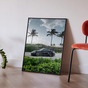 May include: Framed print featuring a black sports car on a coastal road with palm trees under a cloudy sky. The print is leaning against a white wall, next to a red chair and a potted plant.
