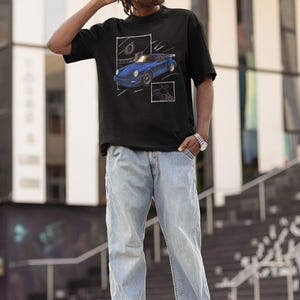 May include: Black t-shirt featuring a blue sports car graphic. The car is depicted in a stylized, artistic manner within a white frame. The person is wearing light blue jeans and white sneakers.