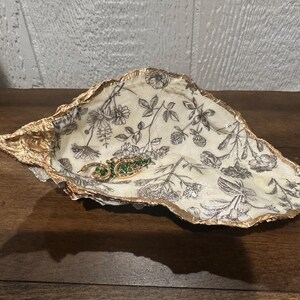 May include: A decorative oyster shell dish with a gold-painted rim. The interior features a cream-colored floral pattern with black botanical illustrations. A gold and green earring rests inside the dish.