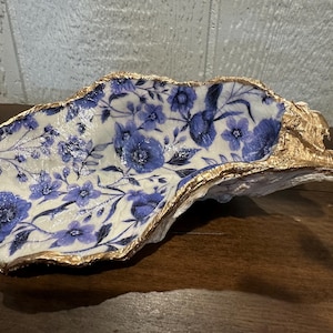 May include: Decorative oyster shell with a floral blue and white pattern. The shell's edge is trimmed with gold paint, adding a touch of elegance. The shell is placed on a wooden surface.
