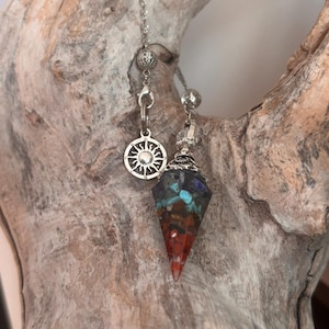 May include: A pendulum with a silver chain and a faceted, multi-coloured crystal point. The crystal features red, orange, blue, and green hues. A silver charm with a sunburst design hangs above the crystal.
