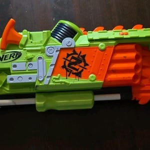 以下が含まれることがあります： ライムグリーンとオレンジ色のNerf Brainsawおもちゃブラスター。前面に回転する鋸刃アタッチメント、黒いトリガー、グレーのラップハンドル、本体側面に黒いZ字型のロゴがプリントされています。