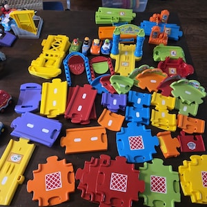 May include: A collection of colorful plastic toy track pieces and accessories, including various straight and curved road segments in red, yellow, blue, green, and orange, along with small toy vehicles, a car wash structure, and a tunnel.