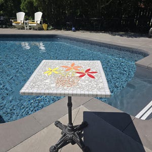 May include: A white mosaic table with a floral design in red, yellow, and orange. The table has a black cast iron base and is situated by a swimming pool.