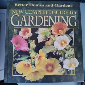 May include: A hardcover book titled Better Homes and Gardens New Complete Guide to Gardening. The dark green cover features a large central photograph of blooming hollyhock flowers in shades of pink, white, orange, and yellow.