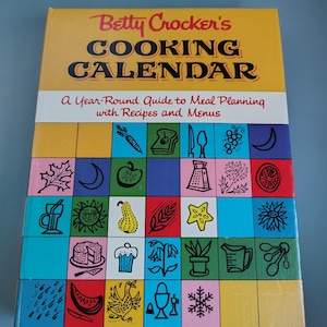 May include: Betty Crocker's Cooking Calendar hardcover book with a yellow top section featuring black text. The cover displays a grid of colorful squares, each containing a black line drawing of various food items, kitchen tools, and seasonal symbols.