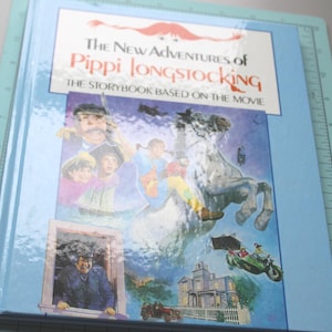 Może przedstawiać: Twarda oprawa książki zatytułowanej "The New Adventures of Pippi Longstocking". Okładka zawiera ilustracje postaci i scen z filmu, w tym Pippi na koniu, domu i samochodzie. Książka ma jasnoniebieską obw&oacute;dkę.