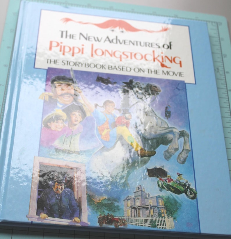 Może przedstawiać: Twarda oprawa książki zatytułowanej "The New Adventures of Pippi Longstocking". Okładka zawiera ilustracje postaci i scen z filmu, w tym Pippi na koniu, domu i samochodzie. Książka ma jasnoniebieską obw&oacute;dkę.